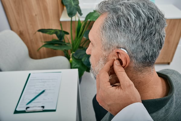 Audiologist Fits A Hearing Aid On Deafness Mature Man Ear While Visit A Hearing Clinic.
